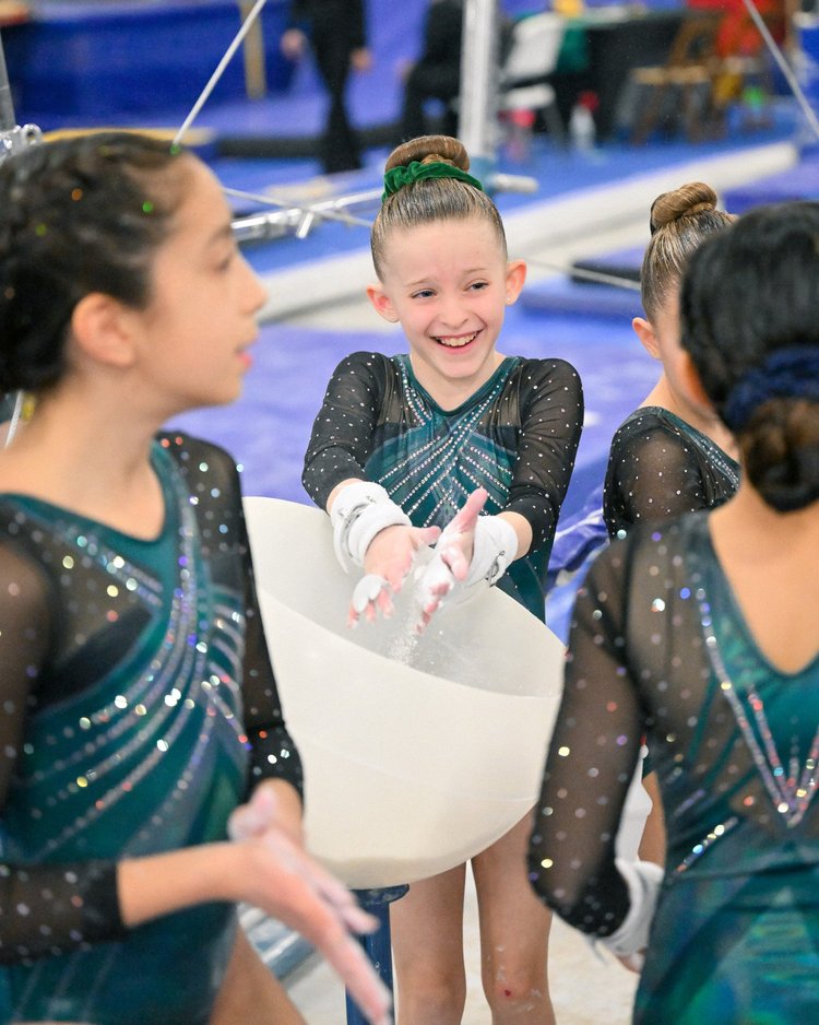 Young gymnasts in teal leotards sharing a candid moment at the chalk bucket